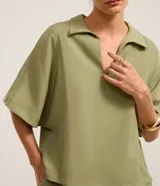 Blusa Cortelle de manga corta amplia y diseño holgado, color verde militar, con cuello solapa y escote en V profundo. Presenta costuras con recortes verticales en los laterales.