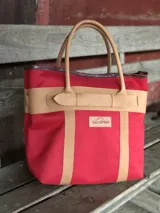 Bolso de playa grande con estampado camuflado en tonos verdes, asas de mano y detalles de tiras en cuero color tostado con hebilla decorativa. Cuenta con un parche de cuero con el logo de la marca en el frente.