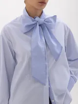 Camisa de poplín con diseño de rayas verticales en tonos azul y blanco. Presenta un cuello prolongado que se anuda en forma de lazo frontal, mangas largas con puños anchos y corte holgado.