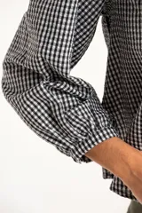 Blusa de manga larga con estampado de cuadros vichy en blanco y negro. Presenta un cuello con volados amplios y un detalle de lazo negro en el centro del escote.