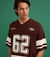 Remera oversized deportiva color marrón con cuello en V, rayas blancas en las mangas y en la parte inferior, y el número "62" estampado en blanco en el frente. Incluye el bordado "Humanity" y un logo de globo terráqueo.