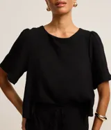 Blusa negra de mangas cortas abullonadas, confeccionada en tejido airflow con textura arrugada y abertura en forma de gota en la espalda.