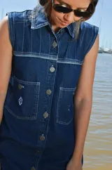 Vestido corto de jean color gris oscuro con efecto acid wash, sin mangas, con cuello camisero y cierre frontal de botones. Incluye dos bolsillos en el pecho con solapa y pespuntes decorativos.