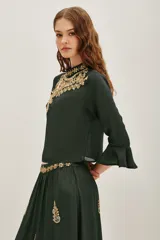 Camisa de viscosa color verde oscuro, con cuello alto y mangas tres cuartos terminadas en volados. Presenta un elaborado bordado artesanal en el pecho con pedrería dorada y detalles decorativos en la cintura.