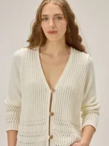 Cardigan de punto color blanco marfil, con escote en V, mangas largas con detalles de tejido calado y cierre frontal de botones con efecto madera.