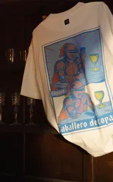 Remera blanca de corte oversize y manga corta, confeccionada en algodón premium. Presenta un estampado gráfico en la espalda con una ilustración estilo cómic de un caballero medieval con una copa y un celular, haciendo referencia a la carta del tarot 'Caballero de copas'.