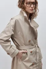 Trench clásico color marrón, de silueta relajada, confeccionado en tejido liviano, con cuello de solapa, doble abotonadura frontal y mangas largas con presillas regulables en los puños.