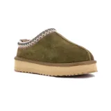 Pantufla tipo zueco de cuero gamuzado color verde oliva, con interior de peluche y suela de plataforma de goma color beige. Presenta un detalle de costura decorativa trenzada en el borde superior.