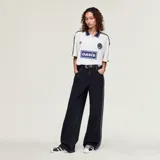 Camiseta Adidas Originals x Oasis de manga corta, color blanco con detalles en azul marino. Presenta cuello polo, las tres tiras características de la marca en los hombros, el logo del trifolio y un estampado frontal con la inscripción "oasis".