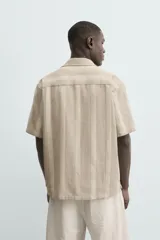 Camisa de calce relajado confeccionada en tejido con mezcla de algodón y lino, con diseño de rayas verticales en tonos beige. Presenta cuello bowling, manga corta, aberturas laterales en el bajo y cierre frontal con botones.