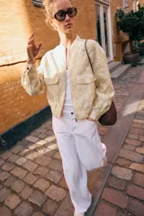 Chaqueta tipo bomber de cuello redondo y manga larga, confeccionada en tejido jacquard con diseño floral en tonos dorados y crema. Presenta cierre frontal con cremallera, bolsillos delanteros de plastrón con solapa y puños y bajo elásticos.
