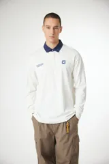 Remera tipo polo de manga larga en color blanco, con cuello en contraste color azul marino. Presenta un pequeño logo cuadrado con una X en el pecho y detalles de texto impreso en el lado derecho.