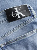 Jeans de mezclilla azul claro, tiro medio, con corte slim que se estrecha hacia el tobillo. Presentan un diseño de cinco bolsillos, bragueta con cremallera y herrajes de Calvin Klein, incluyendo una insignia de cuero en la parte trasera.