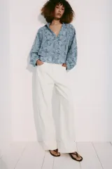 Camisa azul con cuello de solapa, escote en pico y mangas largas con puños abotonados. Presenta un diseño con bordados perforados y patrones geométricos y florales. Cierre frontal con botones.