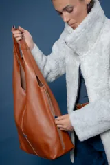 Bolso tipo tote de gamuza en color verde oliva, con diseño minimalista y asa integrada. Presenta una placa metálica con el logo de la marca en la parte inferior central y detalles de textura en el asa.