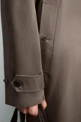 Trench de corte regular confeccionado en tejido técnico italiano repelente al agua. Presenta cuello con solapas de muesca, cierre frontal de botonadura cruzada, cinturón ajustable del mismo tejido, bolsillos de vivo en la cadera, manga larga con puños ajustables y abertura central en la espalda.