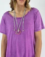 Collar largo con cordones grises y dije de piedra natural irregular color fucsia con borde dorado. Incluye cuentas plateadas y dijes colgantes en forma de corazón.