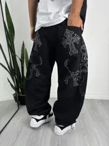 Pantalón de corte baggy color negro con bordados en hilo blanco que presentan diseños de cruces y motivos ornamentales en los bolsillos traseros y las piernas.