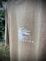 Remera oversize de manga corta, color beige lavado, con estampado circular en la espalda que incluye una palmera y texto que dice "SUNSET", "PALM", "TROPICAL" y "BEACH TRIP".