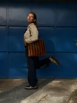 Cartera tipo tote de cuero con diseño de franjas verticales multicolores en tonos tierra, rojo y azul. Cuenta con correas trenzadas de cuero y cierre superior.