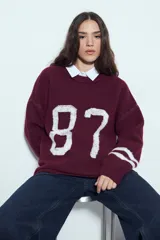 Sweater de punto color blanco con estampado numérico '87' en color negro en el frente y detalles de rayas horizontales en las mangas. Presenta cuello redondo y un corte relajado.