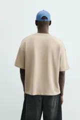 Sudadera de manga corta color beige, corte relajado (relaxed fit) con cuello redondo y número "74" bordado en negro en el frente.