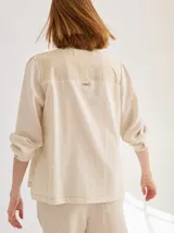 Camisa de corte recto y manga larga, color beige, con canesú trasero de encaje bordado y detalle de placa metálica con logo.