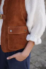 Chaleco de cuero gamuzado color marrón, con cierre frontal mediante dos hebillas metálicas y dos bolsillos frontales con solapa.