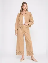 Pantalón de tiro alto y pierna ancha, confeccionado en tela tipo suede color tostado. Presenta bolsillos delanteros, cierre con botón y pasadores en la cintura. El ruedo destaca por un diseño calado con motivos florales.