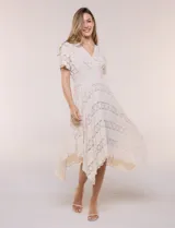 Vestido midi blanco de estilo romántico confeccionado en poliéster con elastano, con detalles de encaje floral. Cuenta con escote cruzado en V, mangas cortas acampanadas, cintura elastizada y falda evasé con terminación asimétrica en picos.