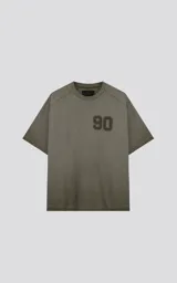 Remera de manga corta, cuello redondo y calce oversize, color verde militar con efecto batik artesanal. Presenta un parche o estampado en el pecho con el número "90".
