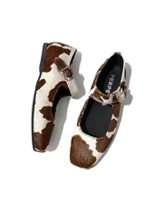 Zapato Mary Janes de cuero con estampado de vaca en tonos marrón y blanco, con correa sobre el empeine y hebilla metálica.