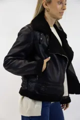 Campera estilo gamulán de cuero sintético negro con forro interno, cuello y puños de corderito negro. Cuenta con cierre frontal cruzado, bolsillos laterales con cierre y trabillas decorativas con ojales metálicos en el cuello.