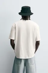 Camiseta de manga corta y cuello redondo, color blanco hueso, con un estampado abstracto multicolor en el pecho que incluye texto en la parte inferior.