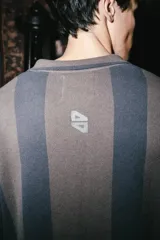 Chomba de manga larga con diseño de rayas verticales en tonos marrón y azul. Presenta cuello polo con cierre de botones y parches decorativos en el pecho.