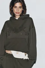 Sudadera con capucha y bolsillo tipo canguro, confeccionada en tejido con efecto lavado. Presenta diseño de estrellas bordadas y aplicación de tachas decorativas en el frente, con terminaciones en rib en puños y ruedo.