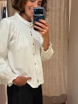 Camisa de algodón color blanco con cuello redondo y detalle de fruncido tipo nido de abeja en el canesú. Presenta cierre frontal con botones decorativos, mangas largas abullonadas con puños elásticos fruncidos y volados en el cuello.