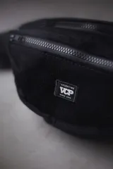 Riñonera de corderoy color negro, con compartimento principal y bolsillo frontal con cierre, tira ajustable con hebilla plástica y detalle de parche bordado con la marca VCP.