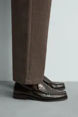 Mocasines de vestir tipo loafer para hombre, confeccionados en cuero marrón oscuro con acabado brillante. Presentan diseño clásico de antifaz en el empeine, costuras visibles en relieve y suela plana con un ligero tacón.