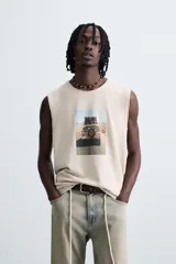 Camiseta tank de punto color beige claro, corte relajado (relaxed fit) y cuello redondo. Presenta un estampado central de un vehículo todoterreno cargado de equipaje conduciendo por una carretera desértica. Los bordes del cuello y las sisas tienen un acabado deshilachado.