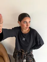 Remera de manga larga color marrón, con cuello redondo y detalle de parche cuadrado con logo bordado en el centro del pecho. Presenta un corte holgado y diseño minimalista.