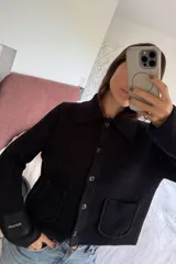 Cardigan negro de punto acanalado con cuello camisero, cierre frontal de botones y dos bolsillos delanteros. Presenta una etiqueta distintiva en la manga.