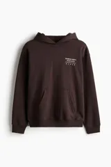 Canguro de mezcla de algodón color marrón oscuro, con capucha cruzada, bolsillo canguro frontal y puños y bajo acanalados. Presenta un estampado pequeño en el pecho con texto y símbolos.