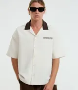Camisa de manga corta, corte boxy, color crema, con cuello inglés y botones frontales. Presenta un bordado pequeño en el pecho izquierdo con texto en color marrón oscuro.