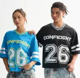 Remera estilo jersey deportiva, color negro, corte boxy y cuello V con ribete blanco. Presenta la palabra "CONFIDENT" estampada en blanco en el pecho y el número "26" grande con efecto desgastado en la parte inferior. Las mangas cortas tienen tres franjas blancas.