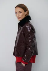 Chaqueta de efecto cuero brillante con acabado charol, cuello con solapas de piel sintética, cierre frontal con botones y bolsillos laterales.