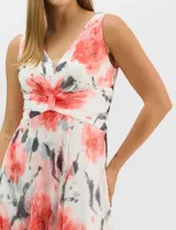 Vestido largo de chifón plisado con estampado floral en tonos rojos y grises sobre fondo blanco. Presenta escote en V pronunciado con panel de microtul, cintura drapeada con nudo central y falda amplia con capa frontal asimétrica. Incluye forro interior y cierre invisible en la espalda.
