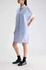 Vestido camisero midi de calce relajado a cuadros en tonos verde y negro, confeccionado en algodón liviano. Presenta cuello clásico en contraste de color blanco, cartera frontal oculta con botones y mangas cortas con vuelta.