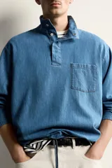 Camisa polo de corte relajado confeccionada en denim ligero de algodón con efecto lavado. Presenta cuello subido, cierre frontal con botonadura oculta por solapa, manga larga, bolsillo de plastrón en el pecho y bajo ajustable con cordón.
