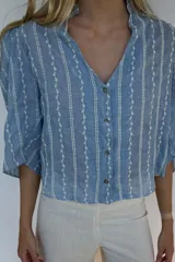 Blusa de algodón liviano en color azul, con diseño de rayas verticales bordadas en blanco. Presenta cuello con volado, escote en V con cierre frontal de botones y mangas 3/4.
